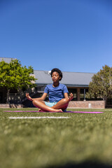 Fototapeta premium African American teen meditating on pink yoga mat on campus field, wearing blue T-shirt and shorts