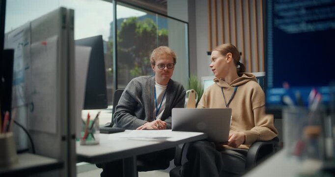 Two Young Software Developers Work and Talk Together on a Laptop in a Modern Tech Office, Discussing App Code, Mobile Ecommerce Features, AI implementation, Cybersecurity as a Video Game Project Team