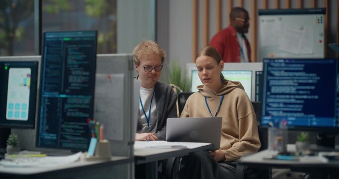  Using Working Computer. Male and Female Software Developers Discuss App Code on Laptop in Tech Office, Surrounded by Coding Monitors, While African American Colleague Reviews Whiteboard Plans Behind.