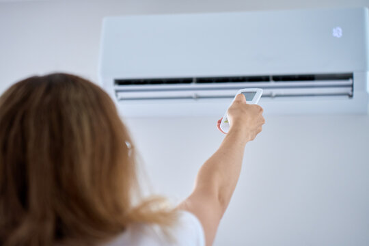 Woman uses a remote control to adjust a wall mounted air conditioner in living room. Concept of cooling, climate control and summer heat.