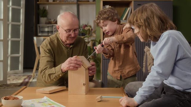 Medium shot of senior Caucasian grandfather helping grandsons assemble wooden birdhouse at home, teaching DIY skills. Video is perfect for family bonding, retirement, and lifestyle ads