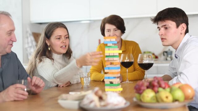 Happy young girl concentrating on removing block from tower while playing jenga, spending free time with boy-friend or husband and parents at light family home 