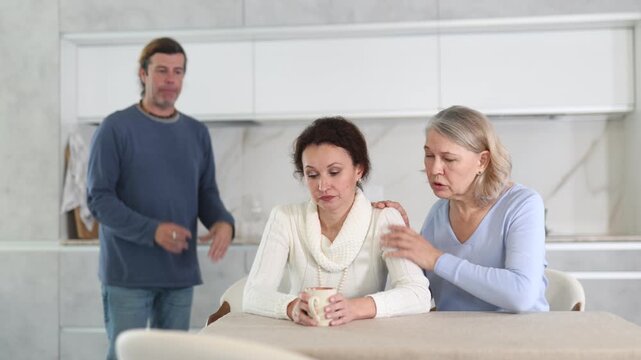 Man is actively quarreling with his thinking wife in kitchen, standing behind her, and elderly mother hugs and calms her daughter