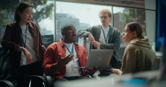 Male Developer Uses Laptop Talks about App Code and Cybersecurity Logic With Three Attentive Colleagues, Sharing Ideas on Ux Design and Data Workflow During a Casual Standup in a Modern Office.