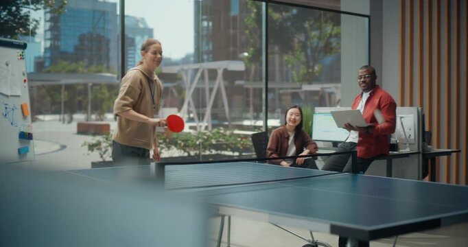 Young White Woman and Male Colleague Play Ping Pong Match in Modern Dev Office While Black Engineer With Laptop Discusses App Code and Cybersecurity With Asian Female Programmer During Team Break.
