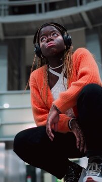 Contemplative female technology user enjoying music beat in headphones while sitting in urban subway station. African American young woman with vitiligo skin listen playlist in earbuds. Slow motion
