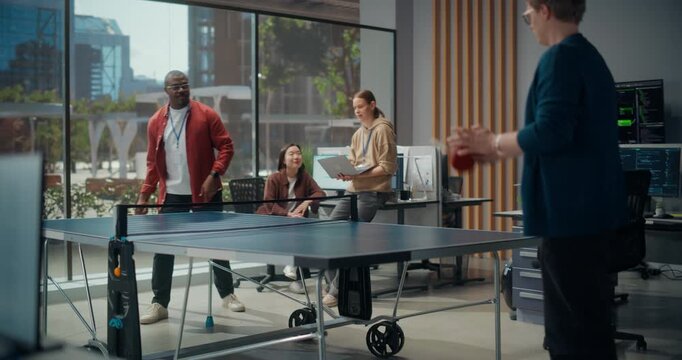 Diverse Software Developers Take Ping Pong Break in Modern Startup Office. Two Male Programmers Play Table Tennis While Female Engineers Watch With Laptop, Code-filled Monitors Glowing Nearby.