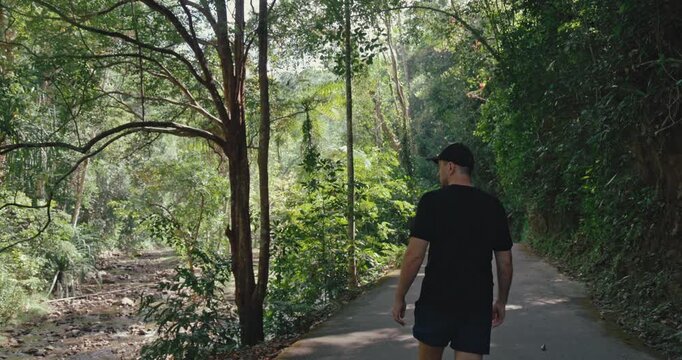 Man in black t-shirt walks along paved road through dense tropical forest. Sunlight filters through trees near rocky riverbed. Active lifestyle and outdoor adventure in nature.
