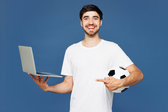 Young shocked IT man fan wear basic white t-shirt cheer up support football sport team use work on laptop pc computer hold in hand soccer ball watch tv live stream isolated on plain blue background