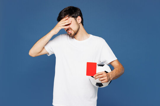 Young man fan wear white t-shirt cheer up support football sport team do facepalm gesture hold soccer ball, red card propose player retire from field watch tv live stream isolated on blue background.
