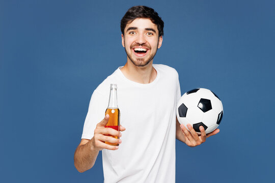Young happy man fan wear basic white t-shirt cheer up support football sport team hold in hand soccer ball watch tv live stream drink beer wink blink eye look camera isolated on plain blue background.