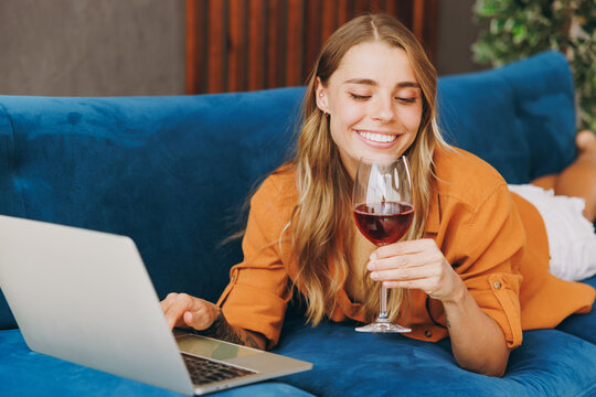 Young satisfied woman wear casual clothes lying on blue sofa watch film or work online on laptop pc computer drink red wine hold glass rest relax indoors flat at home People lifestyle leisure concept