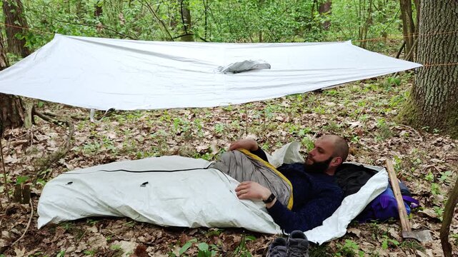 Tyvek bivy bag with a bearded man sleeping under a white poncho tarp in wild forest