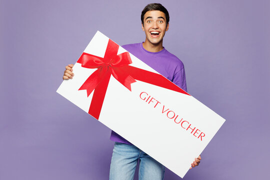 Young surprised shocked man he wears lavender t-shirt casual clothes hold gift certificate coupon voucher card for store isolated on plain pastel purple background studio portrait. Lifestyle concept.