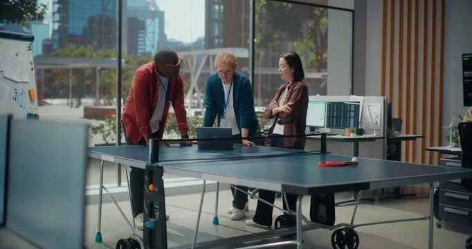 Young Software Engineers meeting Around a Ping Pong Table With a Laptop in a Modern Office, Typing Code and Optimizing App Performance. Team Discuss Cybersecurity and Cloud Solutions.