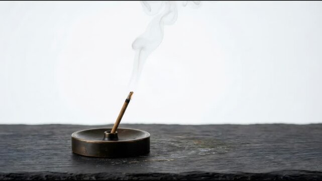Burning incense stick on a small metal holder. Thin trail of white smoke rising into the air. Zen practice for relaxation and meditation in a minimalist space.