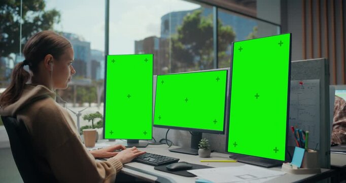 Young Woman Developer Types Code at a Modern Office Desk With Three Green Screen Chroma Key Displays for App Ui Mockup, Cybersecurity Ai Analytics, Cloud Workflow Visualization.