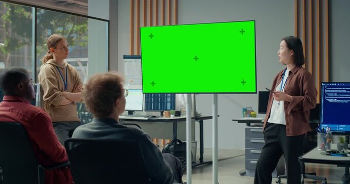 Office Meeting Presentation: Asian Woman Presenter Briefs Software Team in Office, Pointing to Large Green Screen Chroma Key Display. Engineering Ecommerce Platform, Video Game Development Meeting