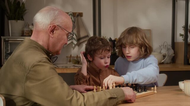 Medium shot of Caucasian senior man and two young grandsons playing chess at home, discussing moves and strategy. Footage ideal for family lifestyle marketing and intergenerational bonding campaigns