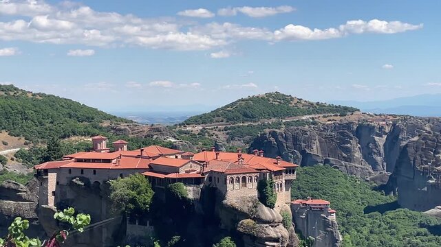 Cinematic slow camera zoom in on the majestic Holy Monastery of Great Meteoron perched high on a steep rocky cliff in Meteora under a beautiful bright blue sky. High quality 4k footage