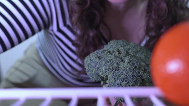 A female picking fresh ingredients from the fridge. POV from inside refrigerator holding broccoli, lemon, celery, lettuce at home.
