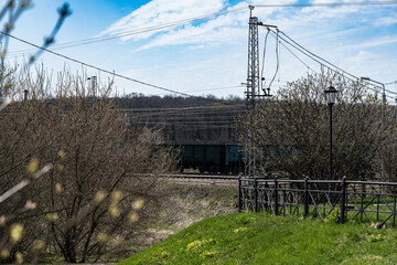 Fototapeta premium Freight train moves through the landscape, partially hidden by budding trees with a bright green foreground. Railway lines and infrastructure are visible, creating a dynamic scene.