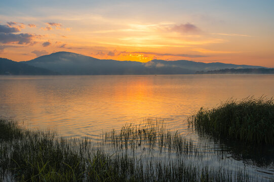 Golden sunrise over a misty mountain lake with grass in the foreground and calm reflective water