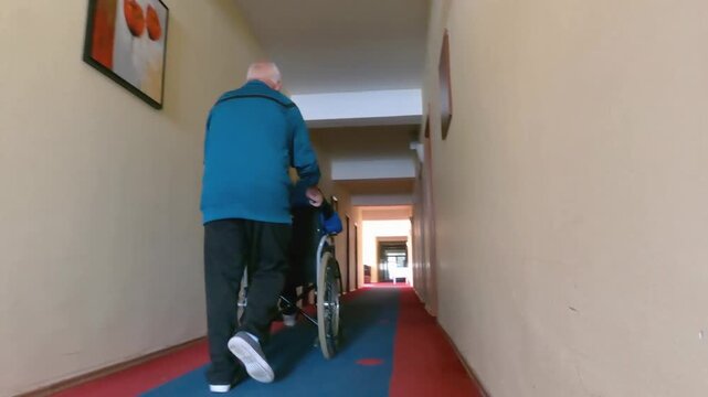 Elderly man assisting a wheelchair user in a hallway of a care facility