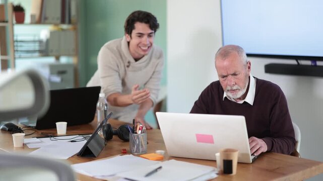 Senior man leaned over to seated guy employee, supervise activities, helps with work and looks at laptop screen. Travel agency, engineering company, notary office or legal advice
