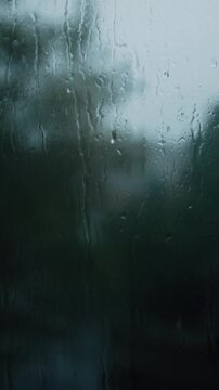 Raindrops stream down a window pane during a heavy downpour, creating a blurry and distorted view of the green trees outside. This moody shot captures the cozy, melancholy atmosphere of a stormy day.