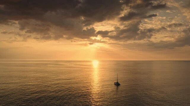 Sailing yacht anchored in calm Andaman Sea at golden hour, orange sun reflecting on tranquil waters under dramatic cloudy sky for a peaceful tropical travel scene. Aerial view drone flight