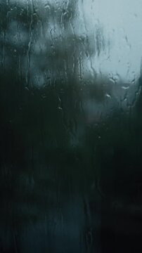 Raindrops stream down a window pane during a heavy downpour, creating a blurry and distorted view of the green trees outside. This moody shot captures the cozy, melancholy atmosphere of a stormy day.