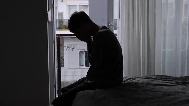 Silhouette man sitting by balcony window, young student on bed with head bowed and hands clasped, soft morning backlight through sheer curtains, urban apartment