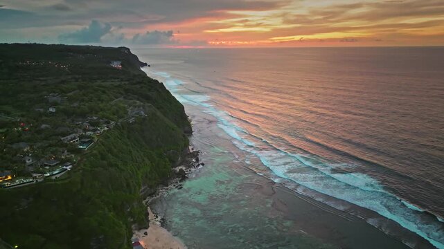 Tropical Bukit Peninsula coastline in evening light, Bali, Indonesia. Lush green cliffs, tranquil turquoise reef water, breaking ocean waves, and vibrant sunset sky. Aerial view drone flight panorama