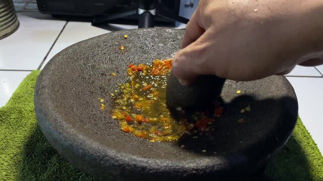 Close-up of hand grinding fresh red chilies in a stone mortar to make sambal paste.  
A detailed stock footage clip showing a person's hand using a stone pestle to grind and crush fresh red hot chilli