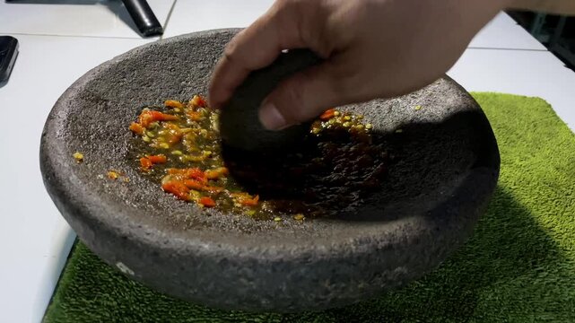 Close-up of hand grinding fresh red chilies in a stone mortar to make sambal paste.  
A detailed stock footage clip showing a person's hand using a stone pestle to grind and crush fresh red hot chilli