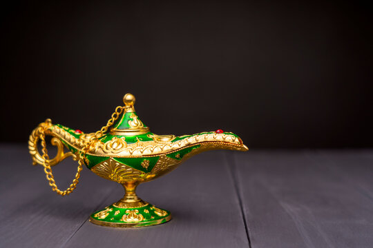 Golden magic lamp with green enamel and red gemstone. Intricate gold filigree decorates the base and handle. Small chain connects the lid to the body. Dark backdrop emphasizes its vibrant colors