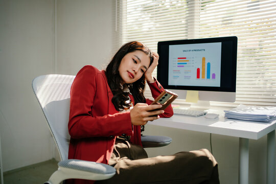 Young Asian businesswoman sitting at her desk, feeling tired and frustrated while looking at her smartphone instead of working