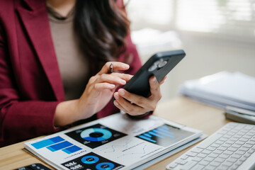Fototapeta premium Professional woman holding a smartphone and pen, analyzing business charts and data on documents and a digital tablet on a desk