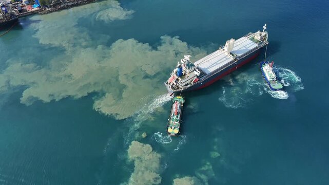 cargo ship assisted by two tugboats while maneuvering in a port, churning up a massive plume of underwater sand and silt into the blue ocean, representing heavy maritime harbor operations.