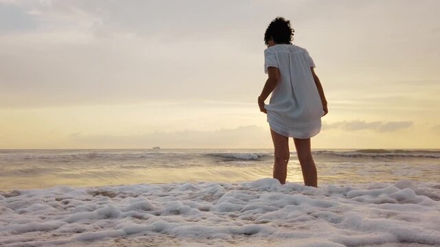 Woman in a white dress stands in the surf as waves splash around her legs