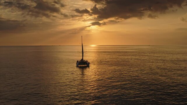 Catamaran sailing on calm ocean waters, carrying people enjoying a beautiful golden hour sunset with sun rays breaking through dramatic clouds above the horizon on Phuket island. Drone flight footage