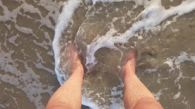 Sea waves splashing submerged tourist feet on sandy beach in ocean with foam