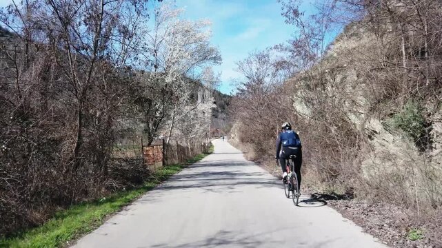 Lone cyclist is pictured on a rural asphalt road, surrounded by scenic landscapes with trees and hilly terrain