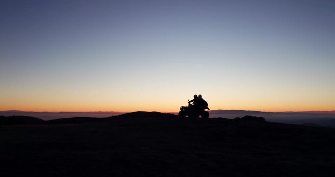 Silhouette explorer couple riding ATV quad bike on mountain summit backlit by at sunset