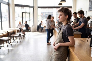 Obraz premium Smiling young barista leaning on a wooden counter in a bright, modern coffee shop, looking thoughtfully away with customers and staff blurred in the background.