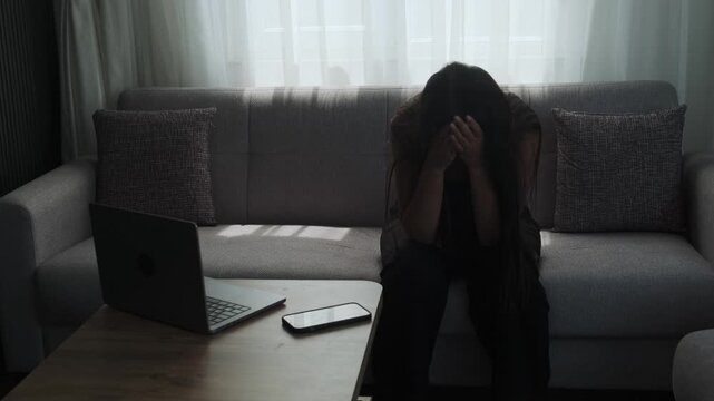 Remote work burnout and digital overload concept with young woman sitting on sofa at home next to laptop and phone, work life imbalance and mental fatigue