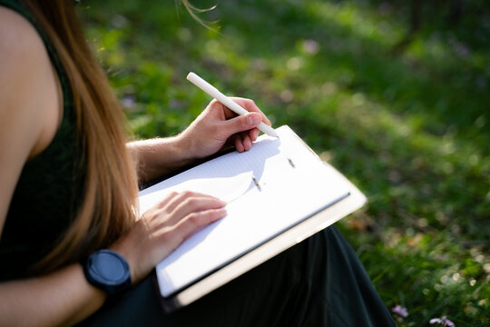 A woman is reflecting on her dreams and recording her goals in a notebook, with a blurry green natural background. The woman improves her self-development and well-being, following a mindful lifestyle