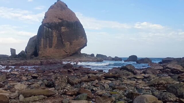 Rocky Tanjung Papuma beach in Jember, Indonesia, featuring a massive sea stack rock formation and crashing ocean waves during low tide.