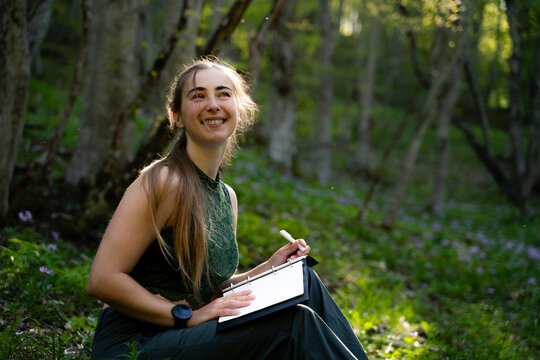 Young cheerful woman relaxing outdoors with her journal and appreciating nature. Concept of self-care, mindfulness and positive emotions.